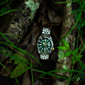 Seiko SSK050K1 GMT watch with green dial and golden bezel sits in a sun-dappled mid-forest, reflecting the sunrise theme of this limited edition timepiece